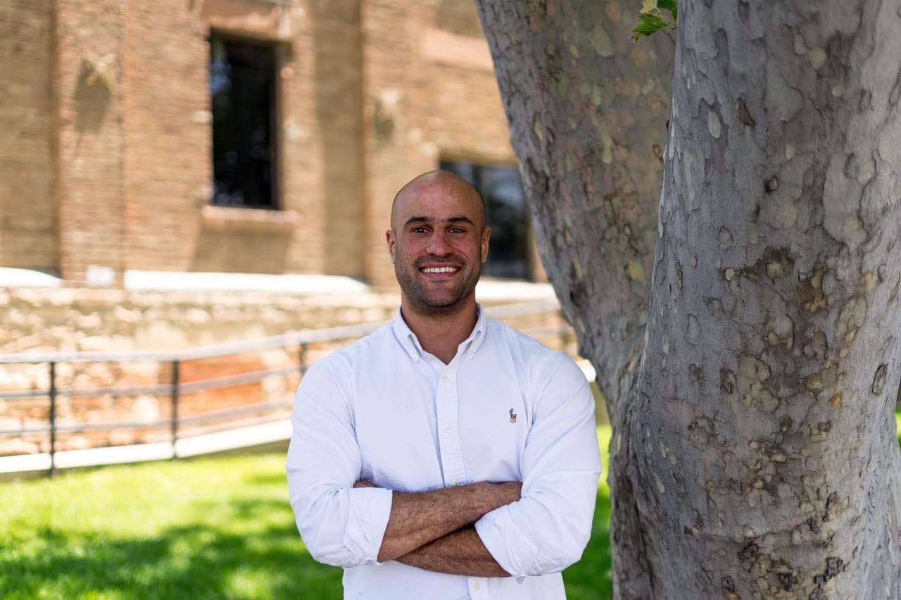 Happy bald man in white shirt, arms crossed, standing by a tree outdoors.