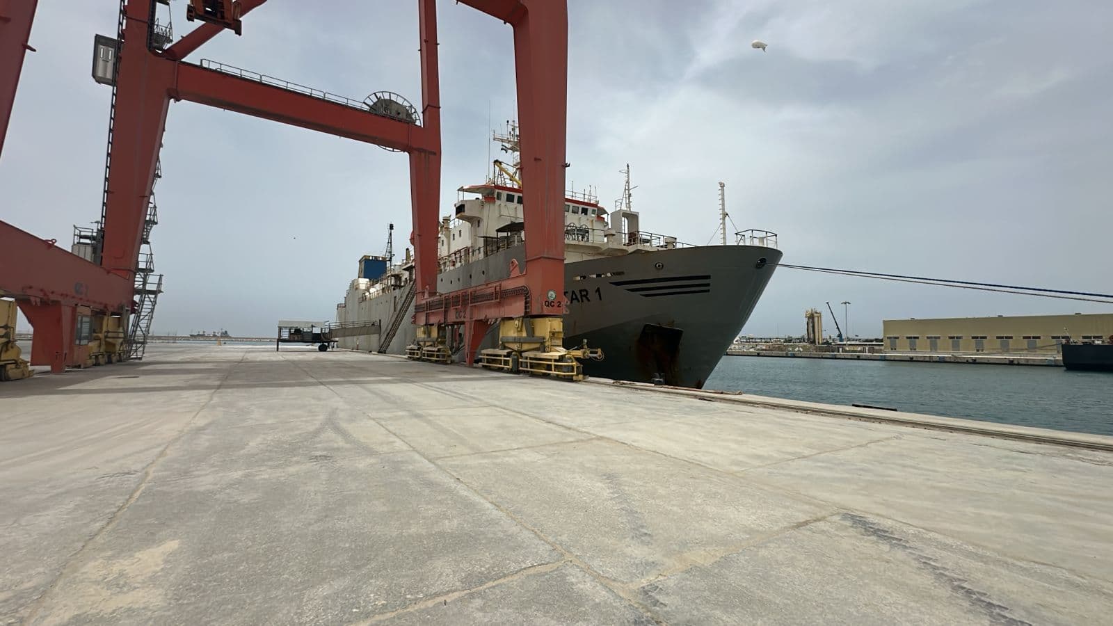 Large cargo ship docked at a concrete port under a massive red gantry crane.
