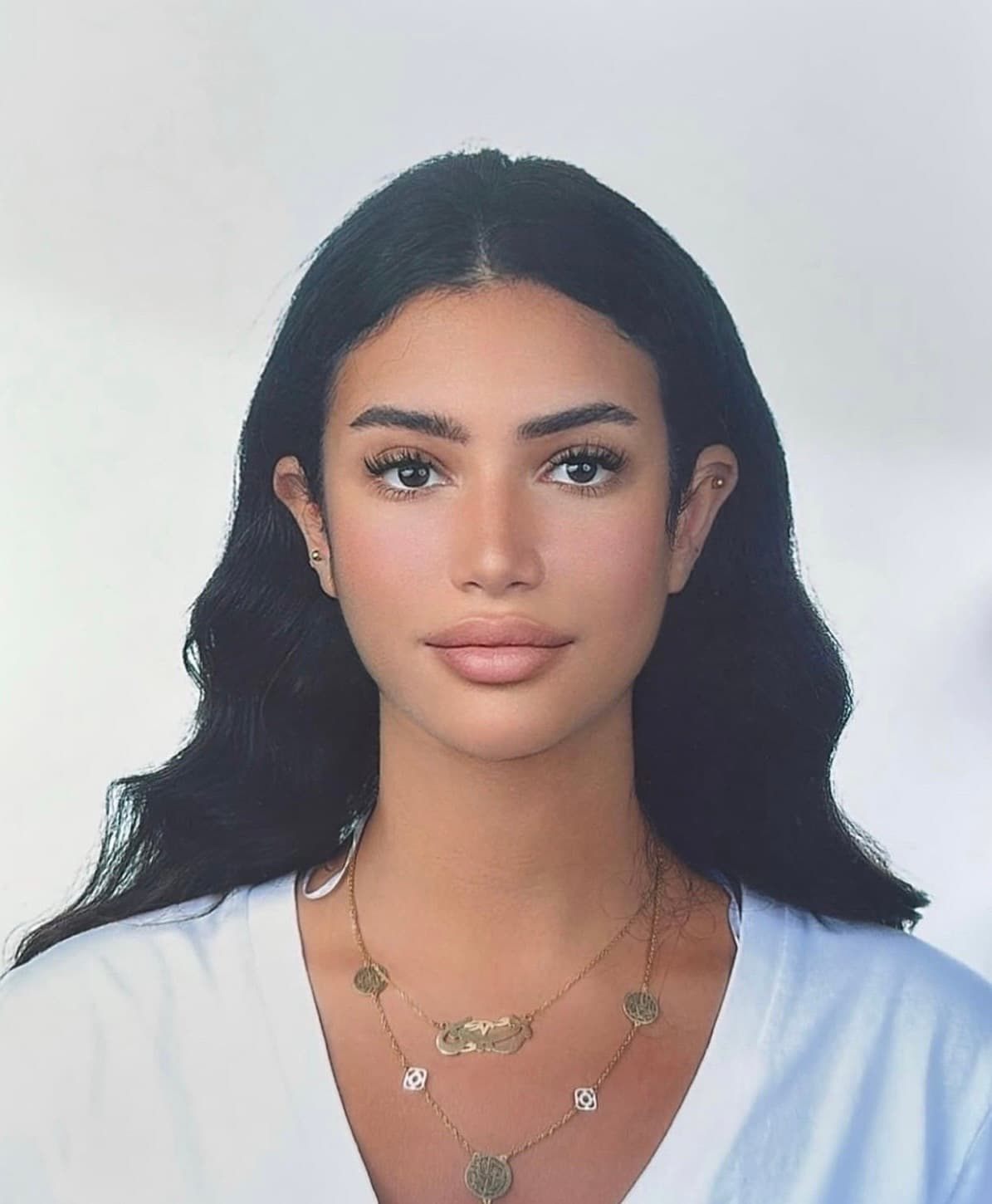 Young woman with long wavy dark hair, wearing gold necklaces against a white background.