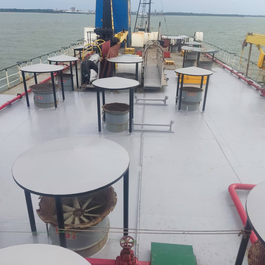 Grey ship deck with round tables, industrial vents, and barrels, overlooking a calm sea.