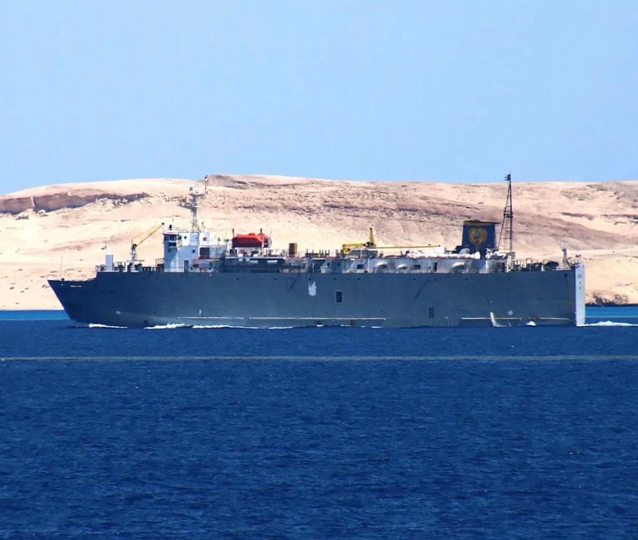Grey transport ship with emblem sailing on blue water past sandy desert hills.